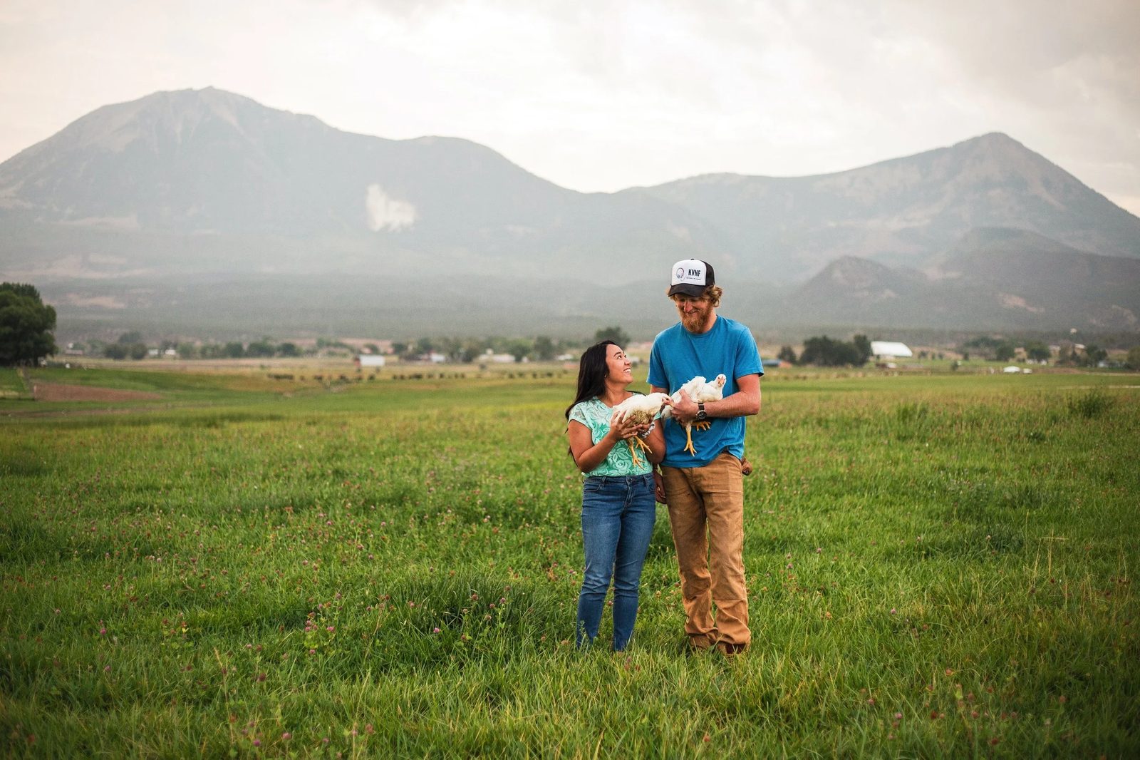 Two farmers holding white broiler chickens in a green pasture, the West Elks above Gray Acres behind them